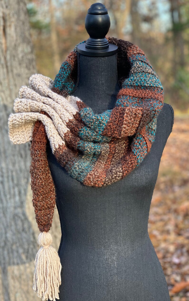 May include: A brown, teal, and cream striped crocheted scarf with a large cream crocheted flower accent and a long cream tassel.