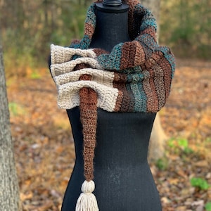 May include: A crocheted scarf with a long tassel in brown, beige, teal, and white colors. The scarf is displayed on a black mannequin.