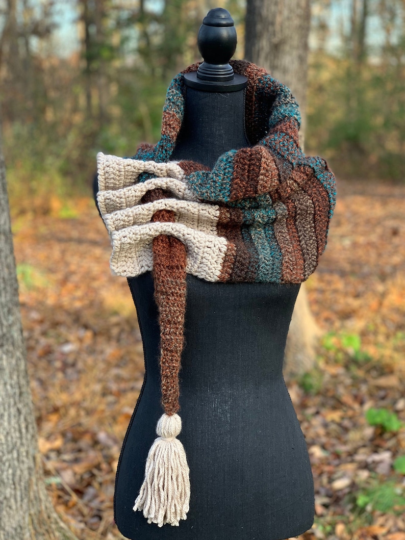 May include: A crocheted scarf with a long tassel in brown, beige, teal, and white colors. The scarf is displayed on a black mannequin.