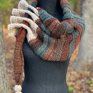 May include: A crocheted scarf with a brown, teal, and cream color scheme. The scarf has a long tassel on one end and is displayed on a black mannequin.