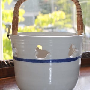 May include: White ceramic bucket with a woven handle. The bucket features duck-shaped cutouts and a blue stripe around the middle. The handle is made of woven light brown material.