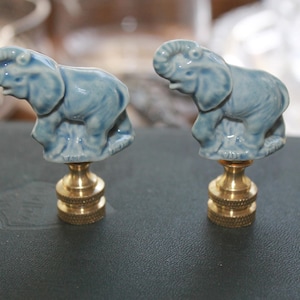 May include: A pair of lamp finials featuring light blue ceramic elephants with raised trunks. Each elephant figure is mounted on a circular, ribbed brass base with a threaded opening, designed to secure a lampshade to a lamp harp.