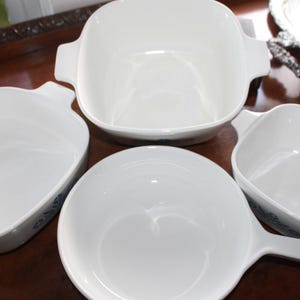 Może przedstawiać: Zestaw białych ceramicznych naczyń kuchennych, w tym duży kwadratowy naczynie z uchwytami, dwa mniejsze kwadratowe naczynia i okrągłe naczynie z uchwytem. Naczynia mają błyszczące wykończenie.
