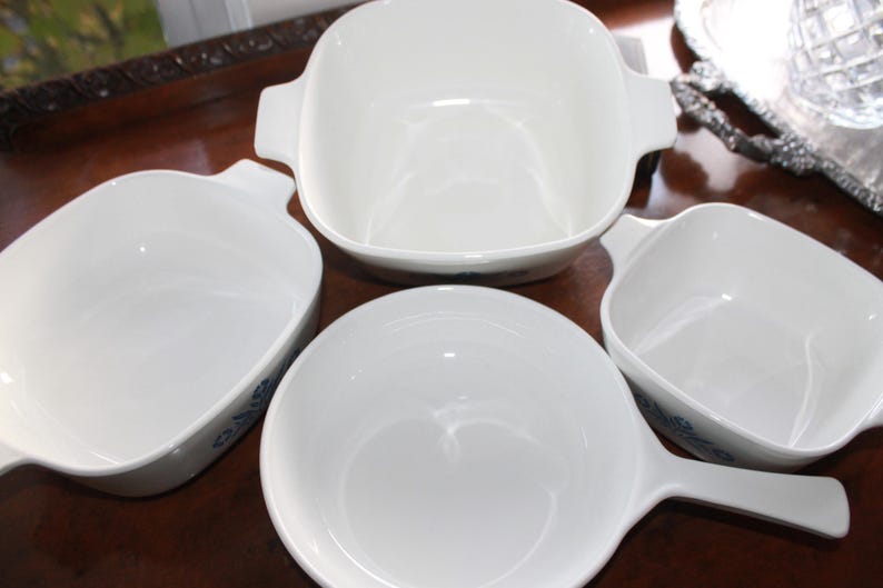 Może przedstawiać: Zestaw białych ceramicznych naczyń kuchennych, w tym duży kwadratowy naczynie z uchwytami, dwa mniejsze kwadratowe naczynia i okrągłe naczynie z uchwytem. Naczynia mają błyszczące wykończenie.