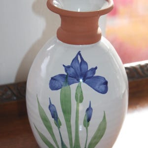May include: White ceramic vase with a blue iris flower design. The vase has a terracotta-colored neck and a wide opening. The iris is painted in shades of blue and green, with leaves and buds. A decorative piece for the home.