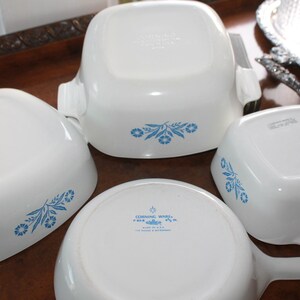Może przedstawiać: Kolekcja białych naczyń CorningWare z niebieskimi kwiatowymi wzorami. Zestaw zawiera naczynie do zapiekania z uchwytami, rondel z uchwytem oraz kilka kwadratowych i prostokątnych naczyń. Naczynia są ułożone na drewnianej powierzchni.