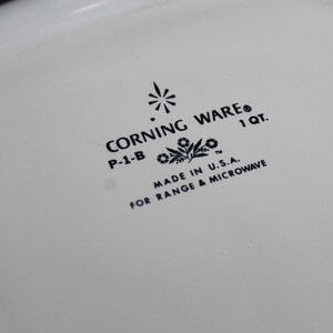 Może przedstawiać: Zbliżenie białego naczynia Corning Ware z czarnym tekstem. Tekst zawiera "CORNING WARE", "P-1-B", "1 QT.", "MADE IN U.S.A." i "FOR RANGE & MICROWAVE". Naczynie jest przeznaczone do kuchenki i kuchenki mikrofalowej.