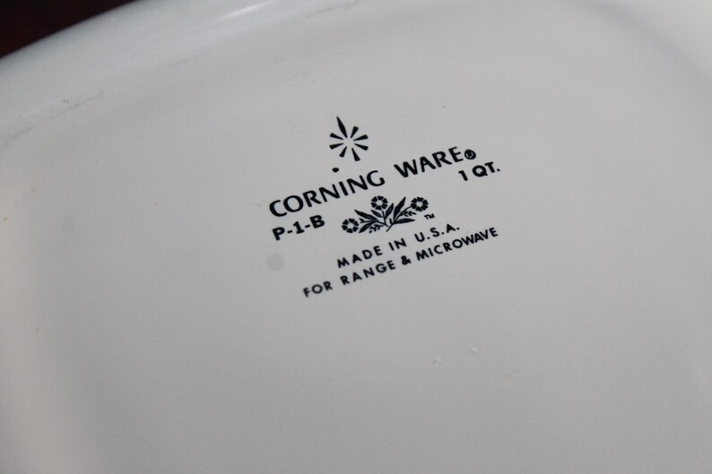 Może przedstawiać: Zbliżenie białego naczynia Corning Ware z czarnym tekstem. Tekst zawiera "CORNING WARE", "P-1-B", "1 QT.", "MADE IN U.S.A." i "FOR RANGE & MICROWAVE". Naczynie jest przeznaczone do kuchenki i kuchenki mikrofalowej.