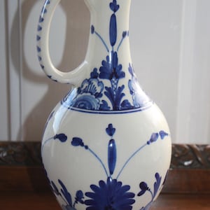 May include: A white ceramic pitcher with a rounded body and a handle. The pitcher is decorated with blue floral patterns and accents. The rim and handle are also detailed with blue designs. The pitcher sits on a blue base.