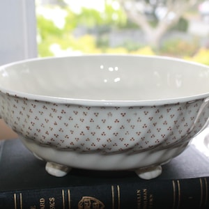 May include: A cream-colored ceramic bowl featuring a repeating pattern of small brown dots arranged in diagonal rows. The bowl has two side handles with green accents, a fluted exterior texture, and stands on three small, rounded feet.