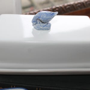 May include: A white ceramic butter dish with a rectangular base and lid. The lid is topped with a small, blue bird figurine. The dish is sitting on a dark surface.