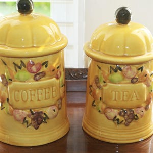 May include: Two yellow ceramic canisters with lids, labeled "COFFEE" and "TEA." Each jar is decorated with a fruit and leaf design. The lids are topped with a dark, round knob. These are kitchen storage containers.