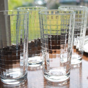 May include: A set of four clear glass tumblers, each featuring a distinct grid pattern of raised squares etched into the exterior surface. The glasses are cylindrical in shape with a flat base and a smooth, unadorned rim at the top opening.