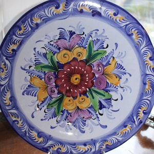 May include: A round, hand-painted ceramic plate with a floral design. The plate features a white center with a bouquet of flowers in shades of red, yellow, and purple, surrounded by green leaves. The rim is blue with a decorative pattern.