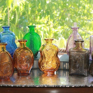 May include: A collection of nine small glass bottles in various colors including amber, blue, green, purple, and smoke gray. Each bottle features embossed figural designs on the front surface and a narrow neck, arranged on a silver-toned metal tray.