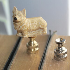 May include: A decorative lamp finial featuring a detailed corgi dog figurine in beige tones. The corgi sits atop a brass base. A second, smaller brass finial is also visible. The items are displayed on a light brown surface.