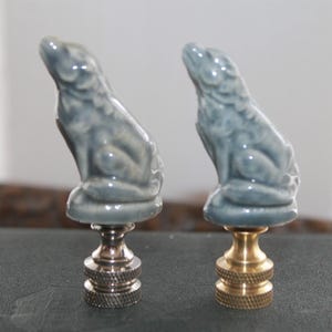 May include: Two ceramic lamp finials in the shape of a sitting dog. The finials are a light blue color and have a glazed finish. One finial is mounted on a silver base, and the other is mounted on a gold base.