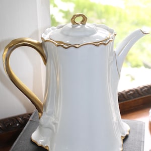 May include: A white porcelain teapot featuring a scalloped edge design, a gold-painted handle, a gold-trimmed spout, and a matching lid with a gold-accented finial. The body of the teapot has a smooth, vertical fluted texture and gold trim.