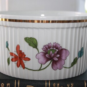 May include: White ceramic serving dish with vertical ribbed design and gold trim. The dish features a floral design with red and purple flowers and green leaves. The dish is sitting on a dark book.