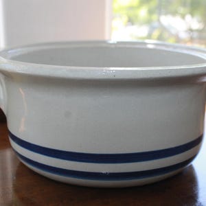 May include: A round, off-white ceramic bowl with two blue stripes around the base and two handles. The bowl is sitting on a wooden surface. The bowl is likely for serving or cooking.