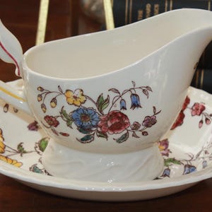 May include: A white porcelain gravy boat with a floral design in shades of pink, blue, yellow, and green. The boat has a handle and a base with a swirl design. It is sitting on a matching saucer.
