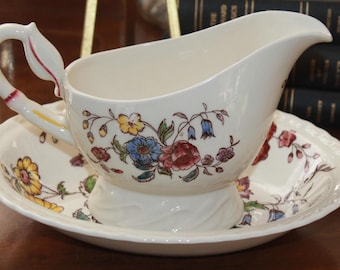 May include: A white porcelain gravy boat with a floral design in shades of pink, blue, yellow, and green. The boat has a handle and a base with a swirl design. It is sitting on a matching saucer.