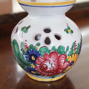 May include: White ceramic vase with floral designs in red, blue, and yellow. The vase has a round body with several circular cutouts and a flared top. A green and white cylinder sits atop the vase. The base and top rim are trimmed with yellow and blue.