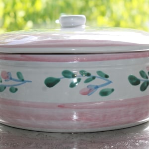 May include: A round white ceramic casserole dish with a lid and two side handles. The exterior features hand-painted horizontal pink stripes and a repeating pattern of green leaves with small pink flower buds encircling the center.