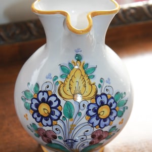 May include: White ceramic pitcher with a yellow rim and base, adorned with hand-painted floral designs in blue, yellow, and green. The pitcher has a bulbous body and a unique spout.