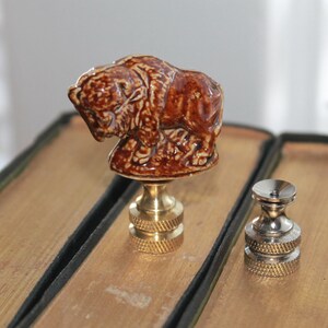 May include: A brown ceramic bison figurine mounted on a gold-toned metal lamp finial base, positioned next to a separate, smaller silver-toned metal lamp finial base with a circular top and a knurled, textured grip on the bottom section.