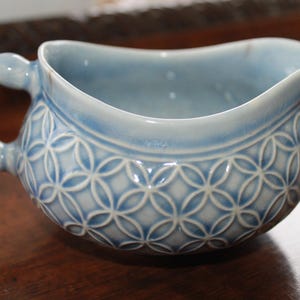 May include: A light blue ceramic creamer with a bamboo-style handle and a spout. The body features a repeating geometric pattern. The creamer is approximately 6 inches long and 4 inches wide.