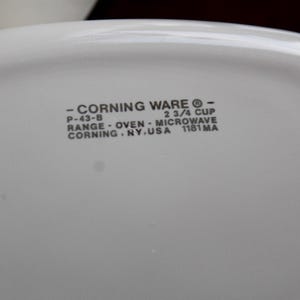 Może przedstawiać: Zbliżenie białego naczynia Corning Ware z zaokrąglonymi rogami. Tekst na naczyniu brzmi "- CORNING WARE - P-43-B 2 3/4 CUP RANGE - OVEN - MICROWAVE CORNING. NY. USA 1181 MA."