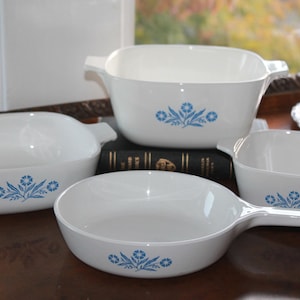 May include: A set of four white ceramic dishes with blue floral designs. The set includes a rectangular baking dish with handles, a square dish, a round pan with a handle, and an oval dish. The dishes are arranged on a dark wooden surface.
