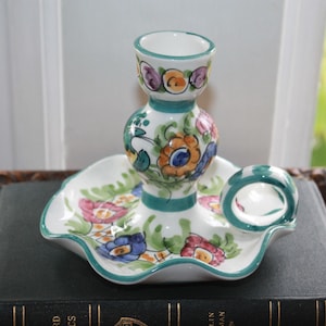 May include: A white ceramic candle holder with a scalloped base and a central column. The holder is decorated with colorful floral designs in pink, blue, and orange, with green accents. A small handle is on the side.