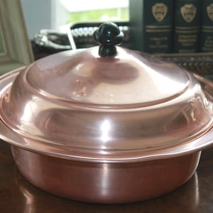 May include: A copper-colored cooking pot with a lid and a black handle. The pot has a round shape with a lid that fits snugly on top. The pot is made of shiny copper and reflects the light.