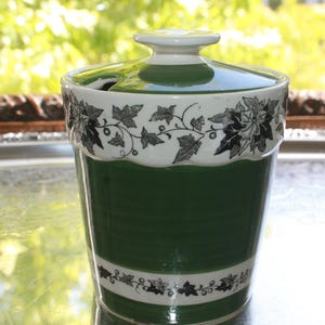 May include: A green and white ceramic jar with a lid. The jar has a cylindrical shape with a white band featuring a black floral design. The lid has a small handle and a matching floral pattern. The jar is likely for storing food items.