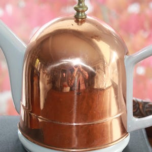 May include: A copper-colored teapot with a white spout, handle, and base. The shiny copper surface reflects the surroundings. The teapot has a brass-colored finial on the lid. The background is a blurred pink and green.