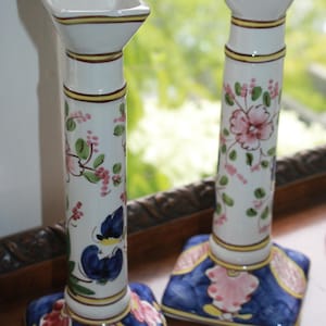 May include: Two white ceramic candlesticks with square bases and tops. The cylindrical stems feature hand-painted floral patterns in pink, green, and blue. The square bases are decorated with deep blue, yellow, and pink accents in a grid-like design.