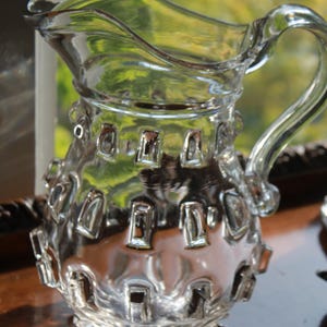 May include: A clear glass pitcher with a decorative design. The pitcher has a handle and a spout for pouring. The body of the pitcher is adorned with rectangular cutouts. The pitcher is sitting on a dark surface.