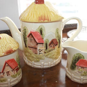 May include: A vintage ceramic tea set with a cottagecore theme. The teapot, creamer, and sugar bowl are decorated with thatched roofs and charming houses. The set is painted in shades of cream, brown, and red.