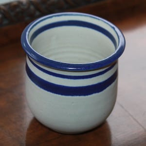 May include: A white ceramic jar with a rounded shape, featuring three horizontal blue stripes and a blue rim. The jar is likely handmade and suitable for kitchen or home decor.