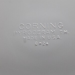 Może przedstawiać: Zbliżenie jasnoszarej powierzchni z wytłoczonym tekstem. Tekst brzmi "CORNING PYROCERAM T.M. MADE IN U.S.A. C-2A". Obraz przedstawia produkt vintage CorningWare.