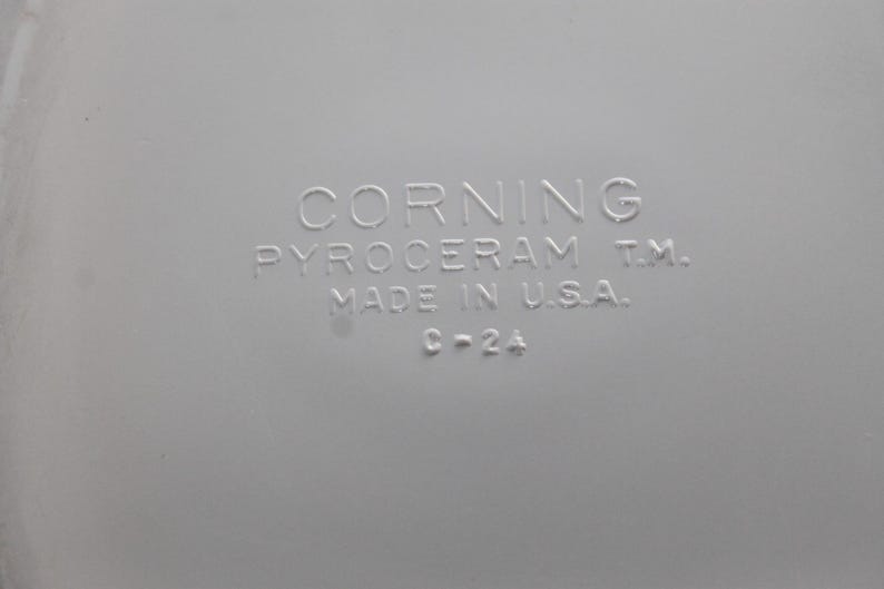 Może przedstawiać: Zbliżenie jasnoszarej powierzchni z wytłoczonym tekstem. Tekst brzmi "CORNING PYROCERAM T.M. MADE IN U.S.A. C-2A". Obraz przedstawia produkt vintage CorningWare.
