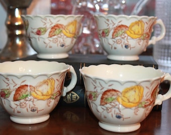 May include: A set of four cream-colored porcelain teacups featuring a scalloped rim and a floral pattern of yellow roses, reddish-brown leaves, and small blue accents. Each cup has a small handle and a pedestal base, arranged in two rows.