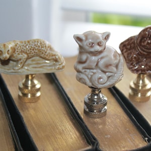 May include: Three decorative lamp finials. One features a leopard with brown spots, another a monkey, and the last an orangutan bust. Each finial is mounted on a brass or silver-colored base. The finials are displayed on a wooden surface.