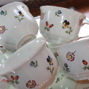 May include: Four white teacups with floral designs. Each cup features a delicate pattern of colorful flowers, including pansies and daisies, with red accents on the handles and rims. The cups are arranged on a reflective surface.
