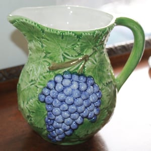 May include: Green ceramic pitcher with a white interior and a large handle. The pitcher is decorated with embossed green leaves and a cluster of blue grapes. The pitcher is a decorative item for the kitchen or dining room.
