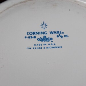 Może przedstawiać: Biały talerz Corning Ware z niebieskim tekstem i kwiatowym wzorem. Tekst brzmi "CORNING WARE", "P-83-B" i "6 1/2 in." (16,5 cm). Zawiera r&oacute;wnież napis "MADE IN U.S.A." i "FOR RANGE & MICROWAVE".
