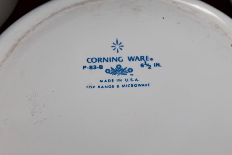 Może przedstawiać: Biały talerz Corning Ware z niebieskim tekstem i kwiatowym wzorem. Tekst brzmi "CORNING WARE", "P-83-B" i "6 1/2 in." (16,5 cm). Zawiera r&oacute;wnież napis "MADE IN U.S.A." i "FOR RANGE & MICROWAVE".