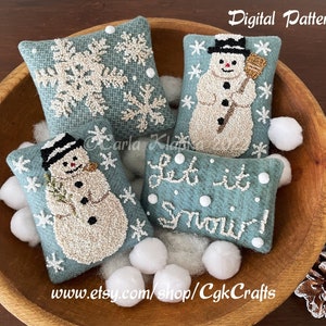 Może przedstawiać: Cztery niebieskie ozdoby filcowe z białymi haftami. Jedna ozdoba przedstawia wzór płatka śniegu, dwie ozdoby przedstawiają wzór bałwana, a jedna ozdoba przedstawia napis "Let it snow!" Ozdoby są prezentowane w drewnianej misce z białymi kulkami z waty.